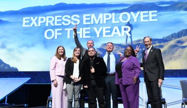 Group of seven people on a stage, one man raising his fist triumphantly, with a backdrop reading "express employee of the year.