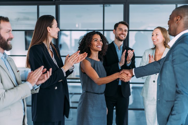 A man and woman are shaking hands while two other men and two other women are looking at them while clapping. Everyone is wearing business attire and the setting appears to be an office lobby.