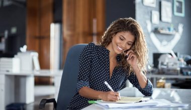 A woman is sitting at a desk and holding a cell phone up to her ear while writing on a notepad.