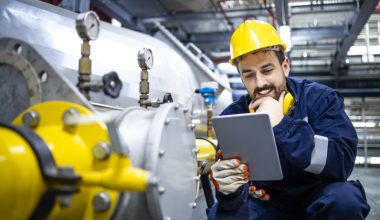 Man in industrial attire using a tablet next to a large pipeline with gauges and valves.