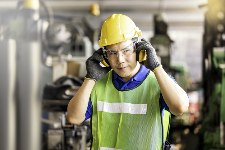 A man wearing a safety vest, hard hat, safety glasses, and protective ear muffs is standing in an industrial plant.