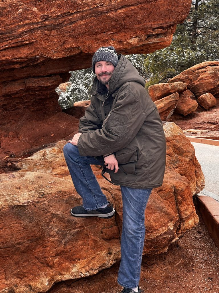 A man in a winter jacket and beanie smiling at the camera while leaning on a large red rock formation.