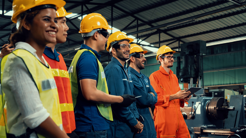A group of workers in safety gear including helmets and reflective vests standing inside an industrial manufacturing plant with machinery in the background. One individual in an orange jumpsuit appears to be holding a tablet.
