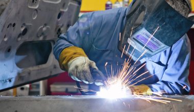 Person welding with protective gear in a workshop, emitting bright light and sparks.