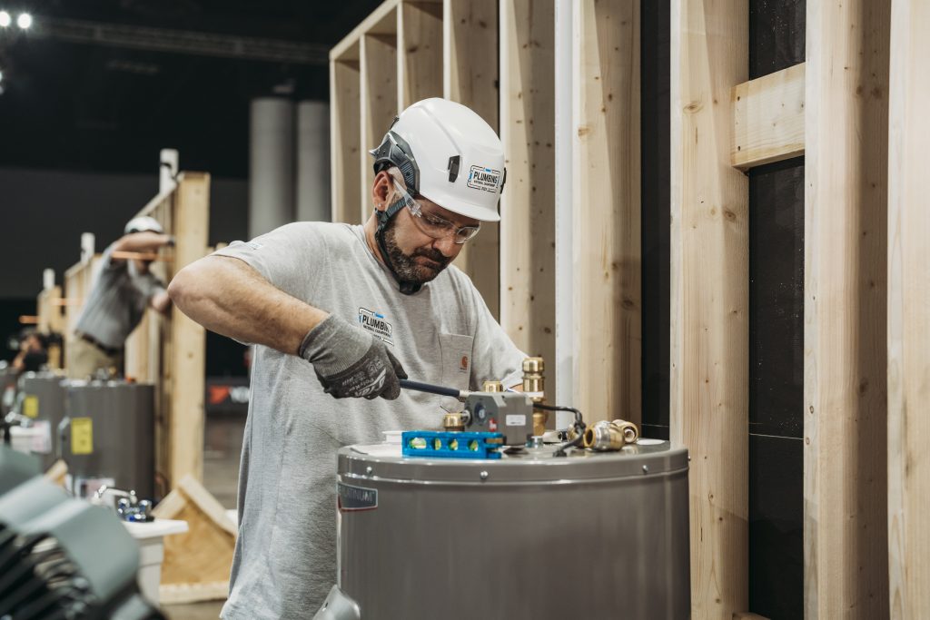 A person wearing a white helmet and gloves is working on a plumbing installation, adjusting equipment on a gray cylindrical tank, with wooden structures in the background.
