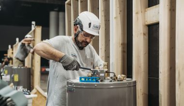 A person wearing a white helmet and gloves is working on a plumbing installation, adjusting equipment on a gray cylindrical tank, with wooden structures in the background.