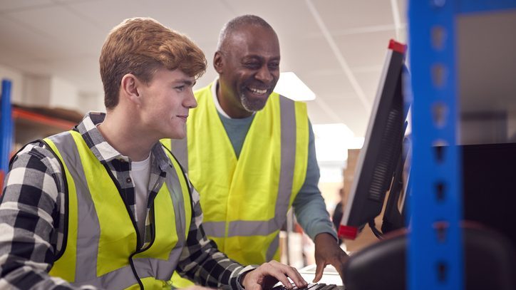 Two men in safety vests, one younger and one older, smiling and looking at a computer screen in a warehouse setting.