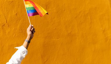 A hand holding a rainbow flag against a bright yellow wall.