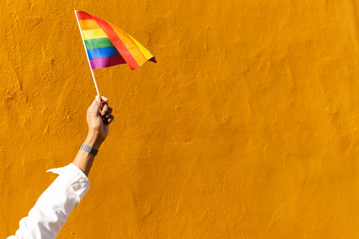 A hand holding a rainbow flag against a bright yellow wall.