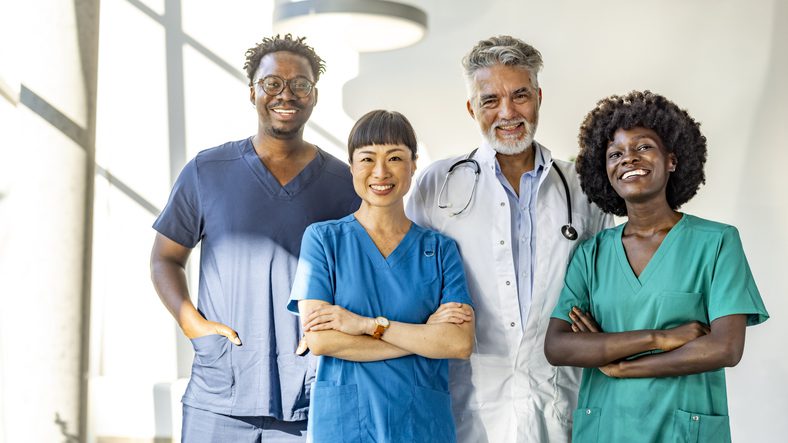 Four healthcare professionals in scrubs and lab coats standing together in a bright, modern indoor space with large windows.