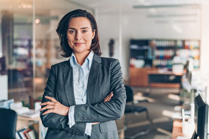 A confident woman in a grey blazer with arms crossed, standing in an office.