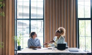 Two women having a discussion at a table in front of large windows in an office space.