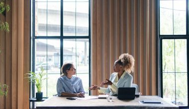 Two women having a discussion at a table in front of large windows in an office space.