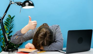 A woman is sitting with her head on her desk beside an open laptop while raising a thumbs up.