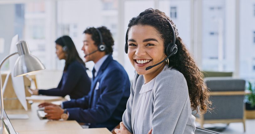 Smiling woman in a headset at an office desk, with two colleagues in the background.