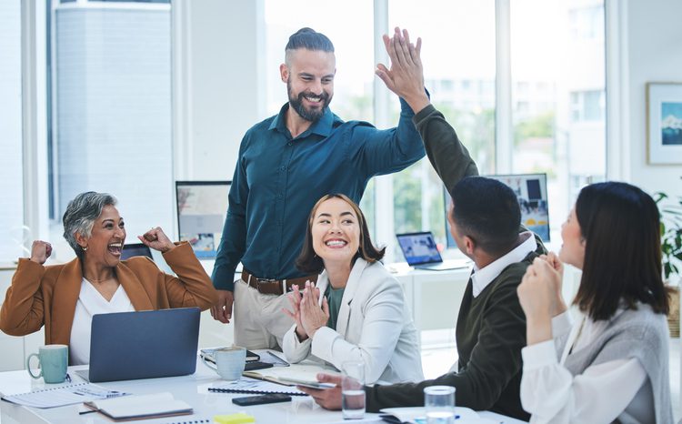 A group of colleagues happily interacting during a meeting, with a man standing and high-fiving a seated man, surrounded by smiling women.