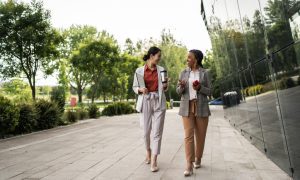 Two women walking and talking on a paved path surrounded by trees and a glass building.