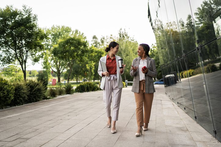 Two women walking and talking on a paved path surrounded by trees and a glass building.