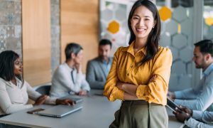 A woman in a yellow blouse stands smiling in front of a conference table with colleagues discussing in the background.