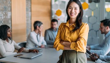 A woman in a yellow blouse stands smiling in front of a conference table with colleagues discussing in the background.