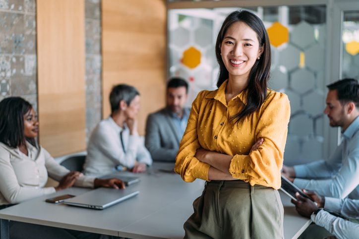 A woman in a yellow blouse stands smiling in front of a conference table with colleagues discussing in the background.