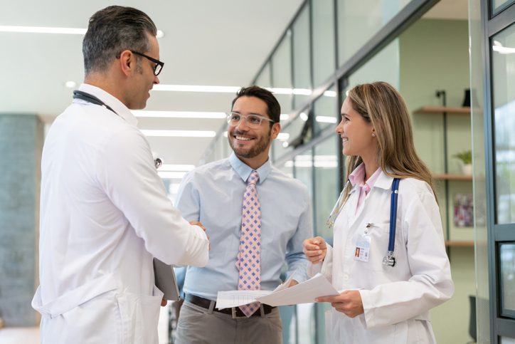 Three professionals in a medical setting, two wearing white coats and one in a blue shirt shaking hands.

