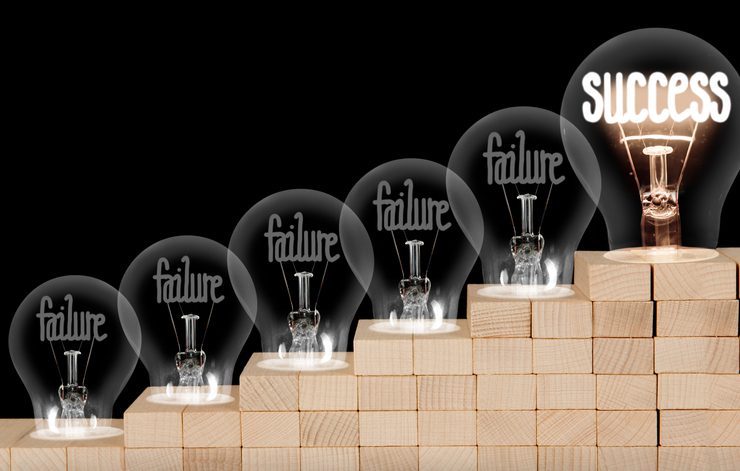 Light bulbs labeled "failure" lead to one labeled "SUCCESS" on ascending wooden steps.