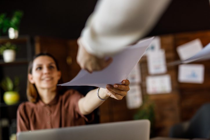 A person smiling and reaching for a document from another individual's hand in an office setting.