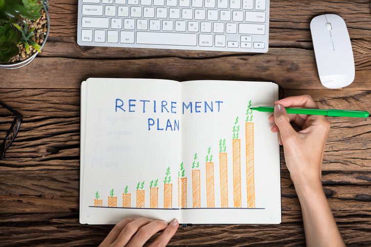 Open notebook with a hand-drawn graph labeled "RETIREMENT PLAN" on a wooden table, with a hand holding a green pen.