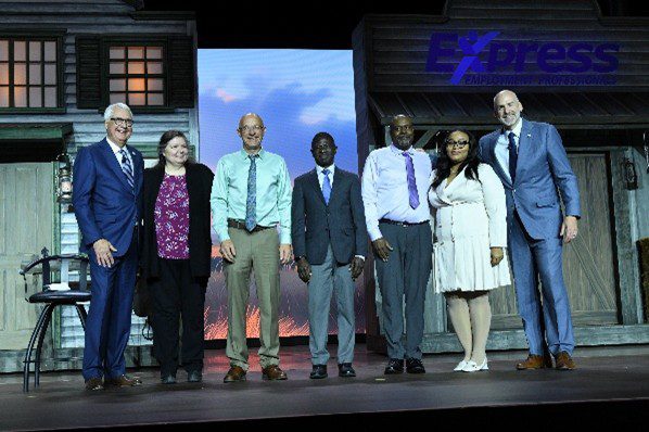 Photo of seven people standing on a stage in front of a backdrop designed like old western-style buildings. The group is dressed in formal and semi-formal attire, including suits, dress shirts, and dresses. The stage background includes wooden doors and windows, and the logo “Express Employment Professionals” is visible in blue at the top right. The lighting highlights the individuals and the set design.