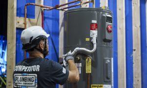 Person in safety gear working on a gray water heater against a blue and wood backdrop.