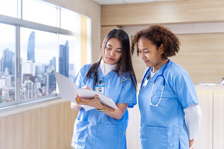 Two healthcare professionals in blue scrubs discussing a document in front of a cityscape window.