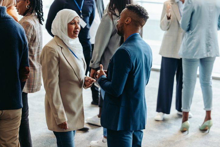 People networking at an event, including a woman in a hijab and a man in a suit talking closely.
