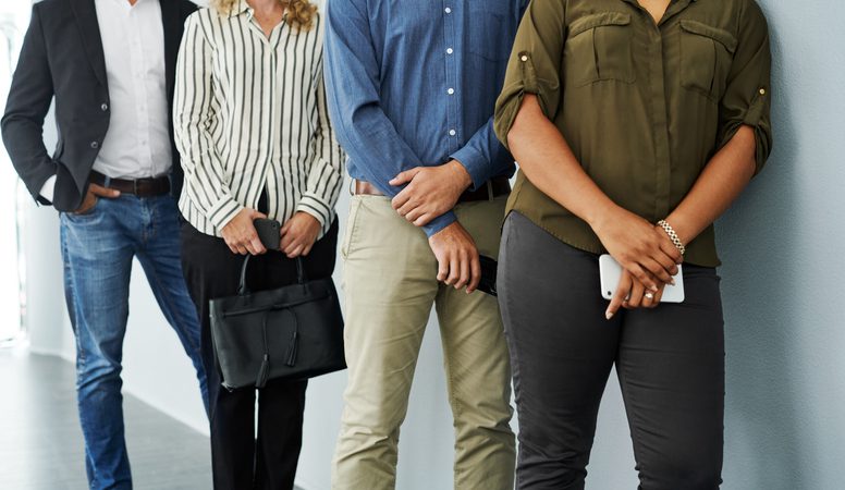 Four people are standing in a line against a wall. The first person on the right is wearing an olive green shirt and holding a smartphone. The second person is wearing a blue shirt and beige pants, with hands clasped in front. The third person is wearing a black and white striped blouse, holding a black handbag. The fourth person on the left is wearing a black blazer over a white shirt and jeans, with hands in pockets.