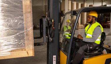 A worker drives a forklift in a warehouse, lifting a wrapped pallet.