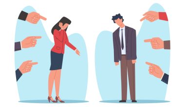 The image shows two individuals standing apart, each surrounded by multiple hands pointing fingers at them. The individual on the left is a person with long hair wearing a red blazer and a dark skirt, covering their face with one hand and lowering the other. The individual on the right is a person with short hair wearing a suit and tie, looking downwards with slumped shoulders.