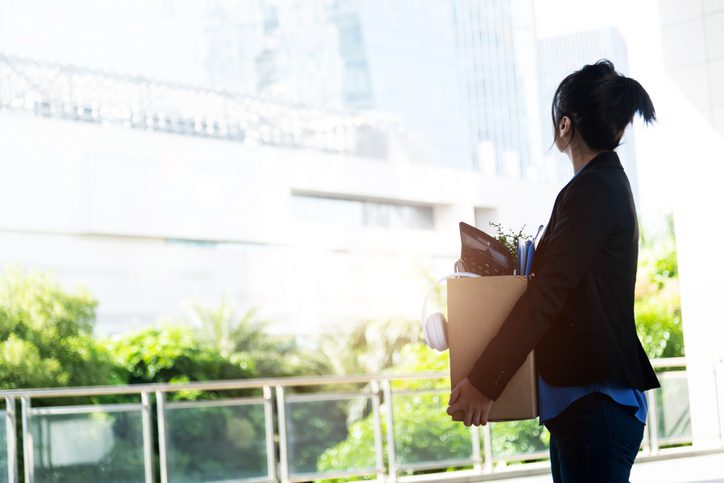 A woman with a ponytail, dressed in a dark blazer and blue shirt, is holding a cardboard box filled with office supplies such as folders, a potted plant, and headphones. She is standing on an outdoor balcony or terrace with greenery in the background and looking towards modern glass buildings.