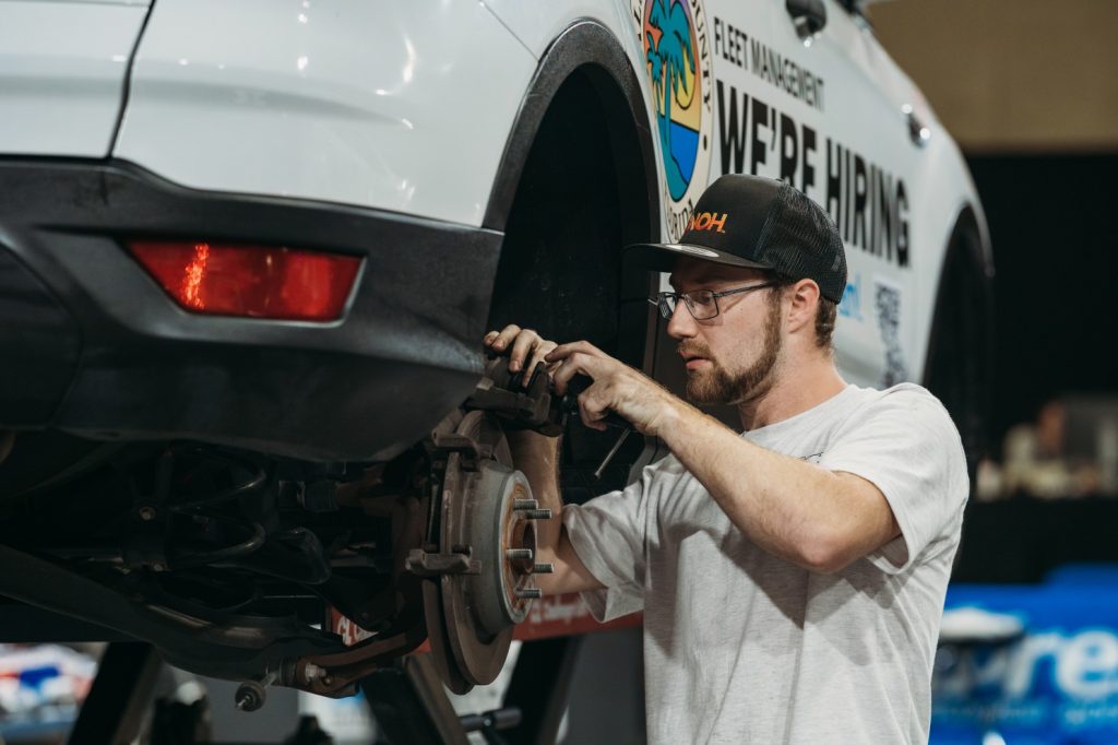 Man working on the rear wheel of a lifted white vehicle with a 'WE'RE HIRING' fleet management sign.