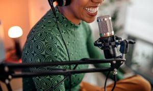 A Black woman in a green patterned shirt wearing headphones and speaking into a microphone in a recording setting.