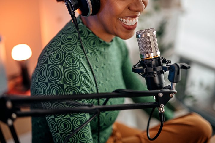 A Black woman in a green patterned shirt wearing headphones and speaking into a microphone in a recording setting.