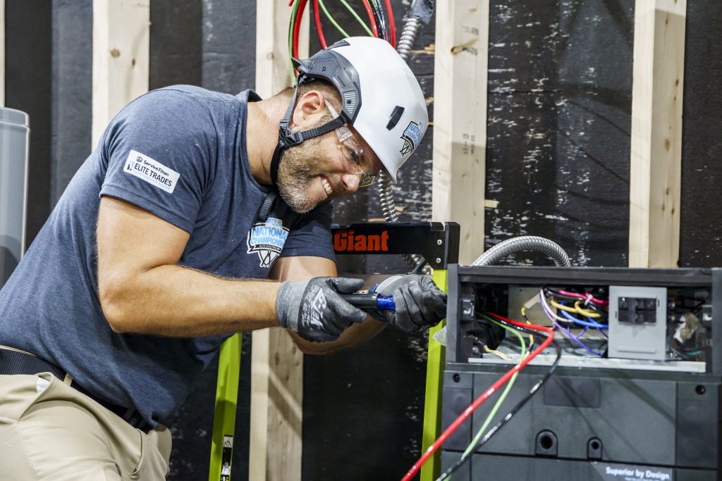 Electrician wearing helmet and gloves working on wiring inside an electrical panel, with visible colored wires
