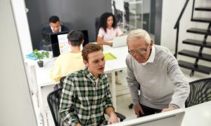 Multigenerational team working in a modern office. An older employee assists a younger colleague at a computer, promoting collaboration and mentorship.