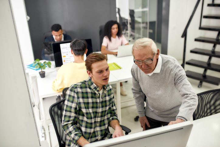 Multigenerational team working in a modern office. An older employee assists a younger colleague at a computer, promoting collaboration and mentorship.