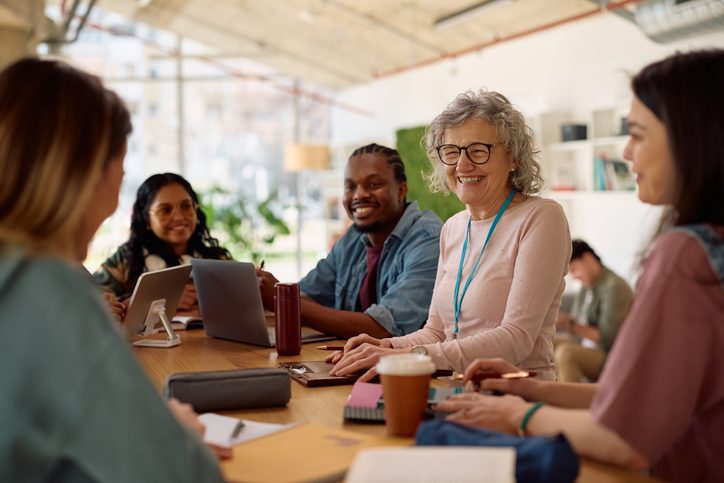 Group of diverse professionals engaged in a collaborative meeting around a table in a modern, well-lit office space. Participants are actively conversing and using laptops and tablets, with notebooks, coffee cups, and office supplies scattered across the table. Large windows provide natural light, enhancing the open and contemporary workspace.