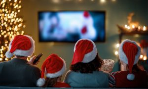 Four people wearing red and white Santa hats sit closely together on a couch, viewed from behind, watching a holiday movie on TV. One holds a remote control, another a bowl of popcorn. A lit Christmas tree and festive lights decorate the cozy room, creating a warm and cheerful holiday atmosphere.