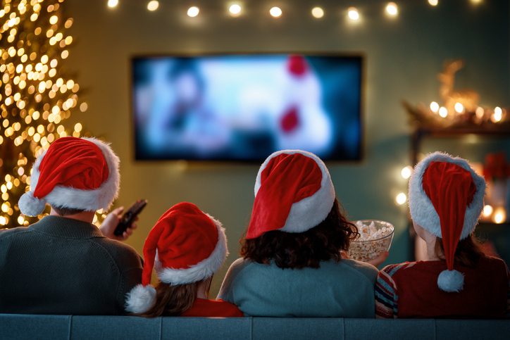 Four people wearing red and white Santa hats sit closely together on a couch, viewed from behind, watching a holiday movie on TV. One holds a remote control, another a bowl of popcorn. A lit Christmas tree and festive lights decorate the cozy room, creating a warm and cheerful holiday atmosphere.
