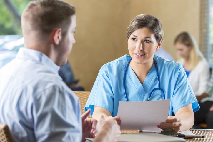 Healthcare worker in scrubs chats with someone at a table in a busy indoor space.