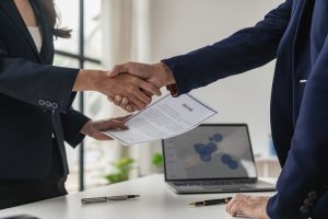 Two people in business attire shaking hands across a desk, one holding a document labeled ‘Resume.’ A laptop displaying a chart and a pen are visible on the desk.