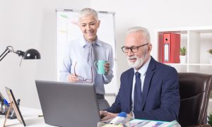 Two older adults in a professional office setting smiling at a laptop screen. One stands holding a green coffee mug and red eyeglasses, while the other sits in a dark blue suit and tie. The desk features a laptop, lamp, framed photo, pen holder, and documents with charts. A flip chart, red binder, and potted plant are visible in the background.