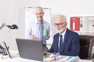 Two older adults in a professional office setting smiling at a laptop screen. One stands holding a green coffee mug and red eyeglasses, while the other sits in a dark blue suit and tie. The desk features a laptop, lamp, framed photo, pen holder, and documents with charts. A flip chart, red binder, and potted plant are visible in the background.